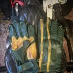 Hold Fast 1971 Le Mans Handschuhe aus patiniertem grünem Leder, von der Handfläche und vom Handrücken auf einem gepolsterten Motorradsitz gesehen.
