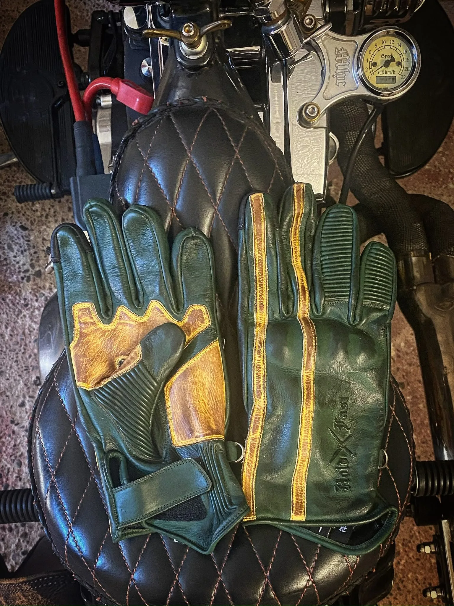 Hold Fast 1971 Le Mans Handschuhe aus patiniertem grünem Leder, von der Handfläche und vom Handrücken auf einem gepolsterten Motorradsitz gesehen.