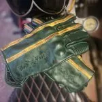 Detailansicht der grünen Lederhandschuhe „Hold Fast Le Mans“ mit ockerfarbenen Streifen und geprägtem Logo, die auf einem Sattel liegen.