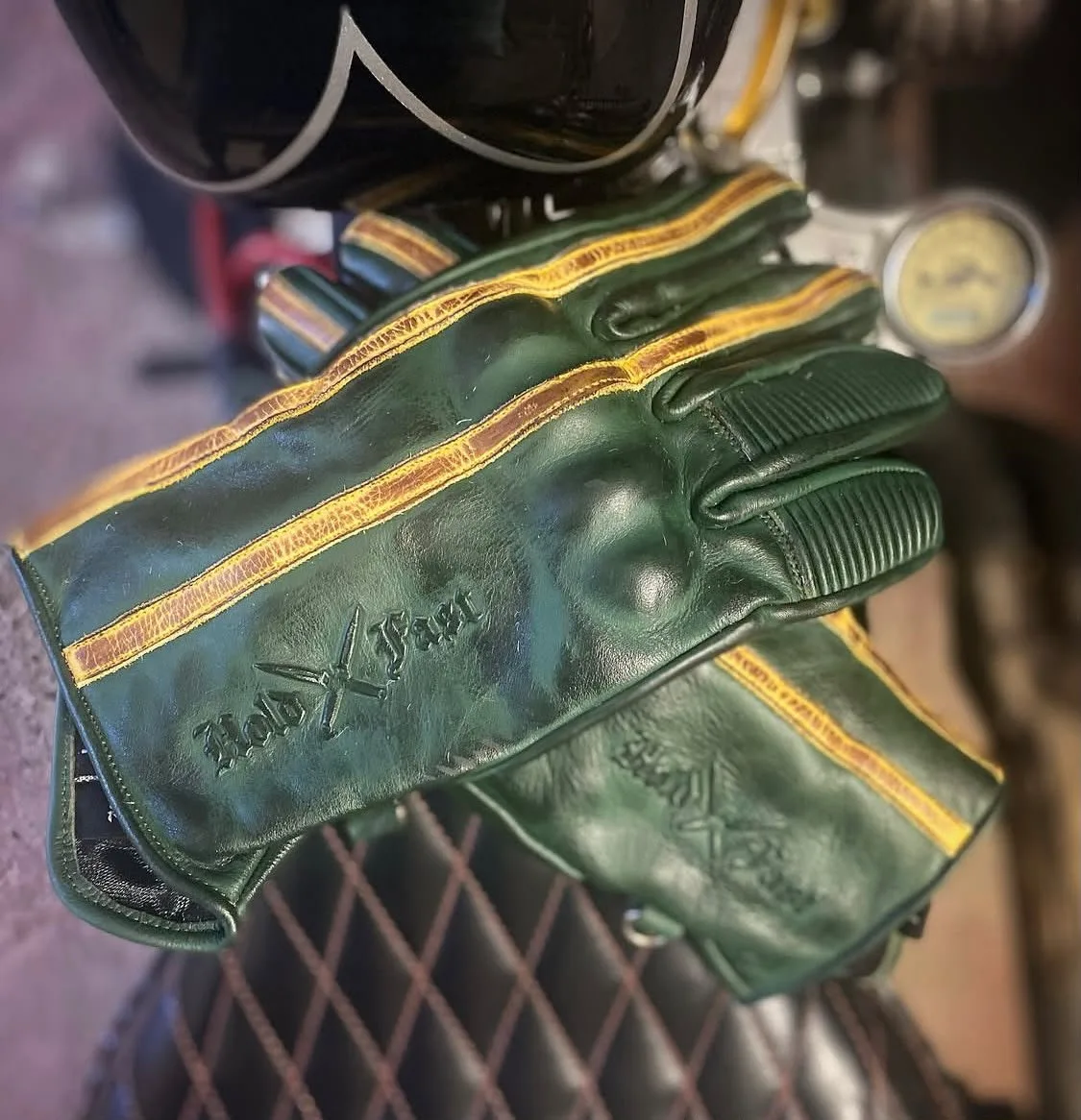 Detailansicht der grünen Lederhandschuhe „Hold Fast Le Mans“ mit ockerfarbenen Streifen und geprägtem Logo, die auf einem Sattel liegen.